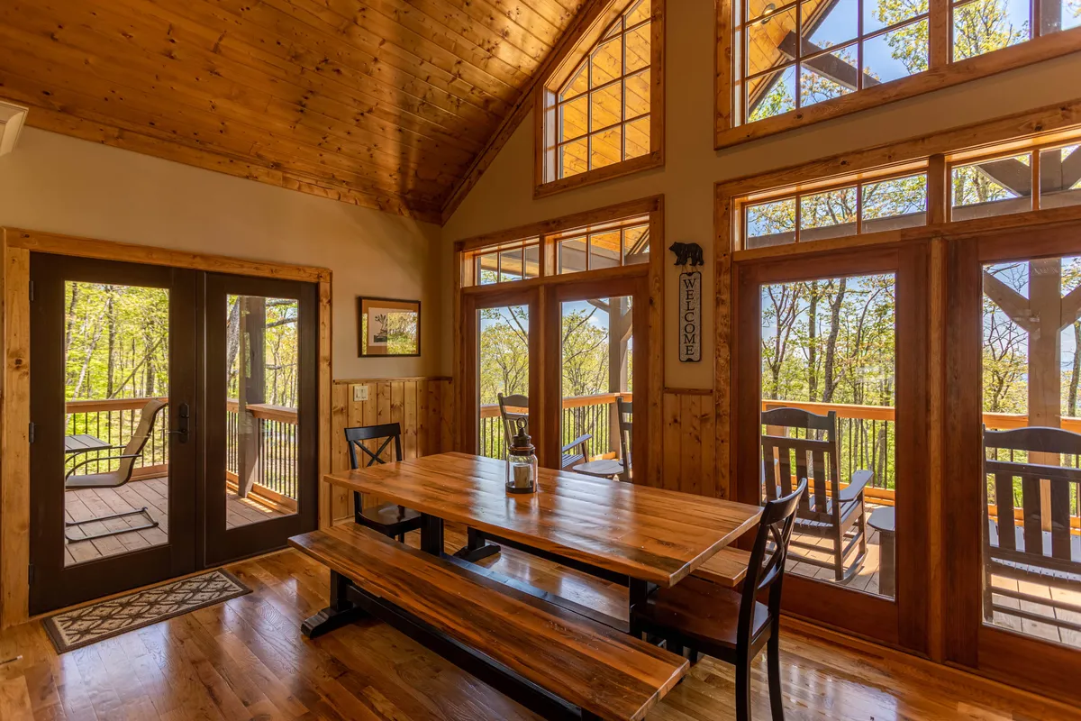 A large wooden dining table is set beside expansive windows and under a ceiling of wooden beams, with sliding glass doors offering more light and views. The space is perfect for group meals, conversations, and enjoying the natural surroundings from inside.