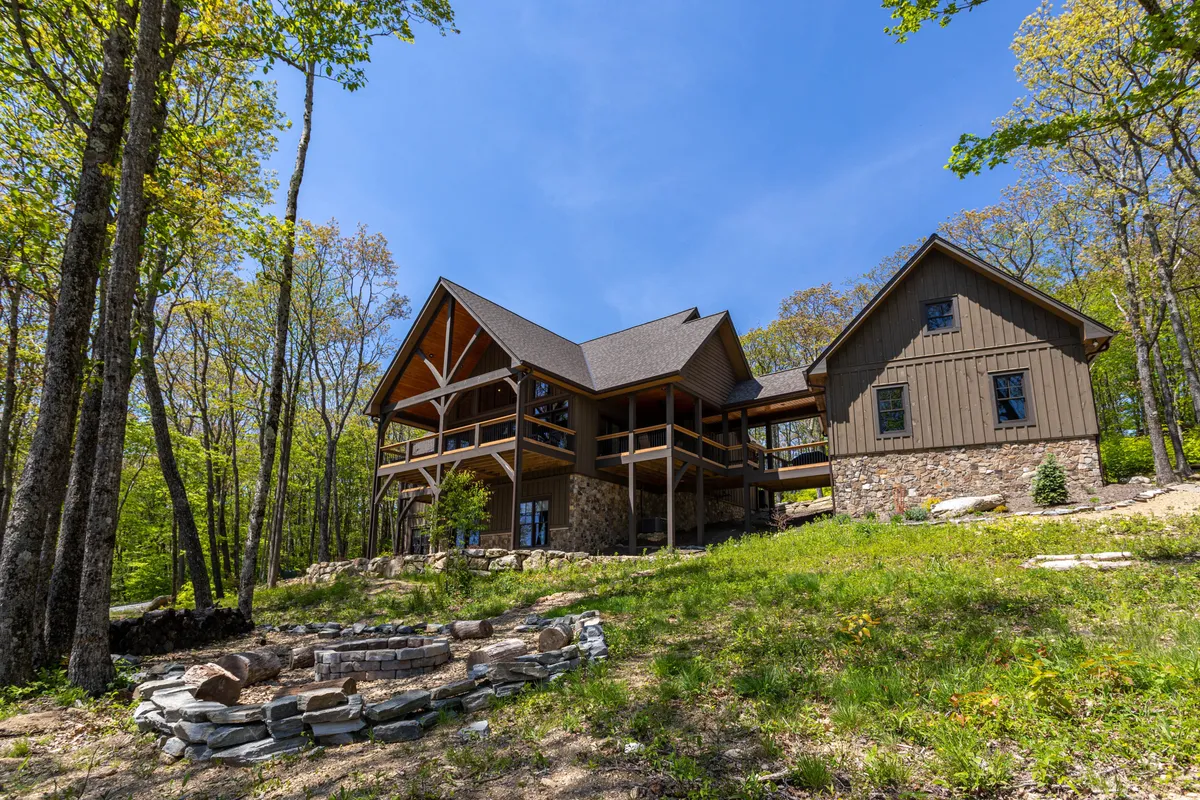 The image shows a spacious, rustic wooden cabin perched on a ridgetop, surrounded by tall trees. The stone patio features a welcoming fire pit, perfect for group gatherings. The porch offers sweeping sunset views over the Blue Ridge Mountains, inviting guests to relax and soak in the tranquil setting.