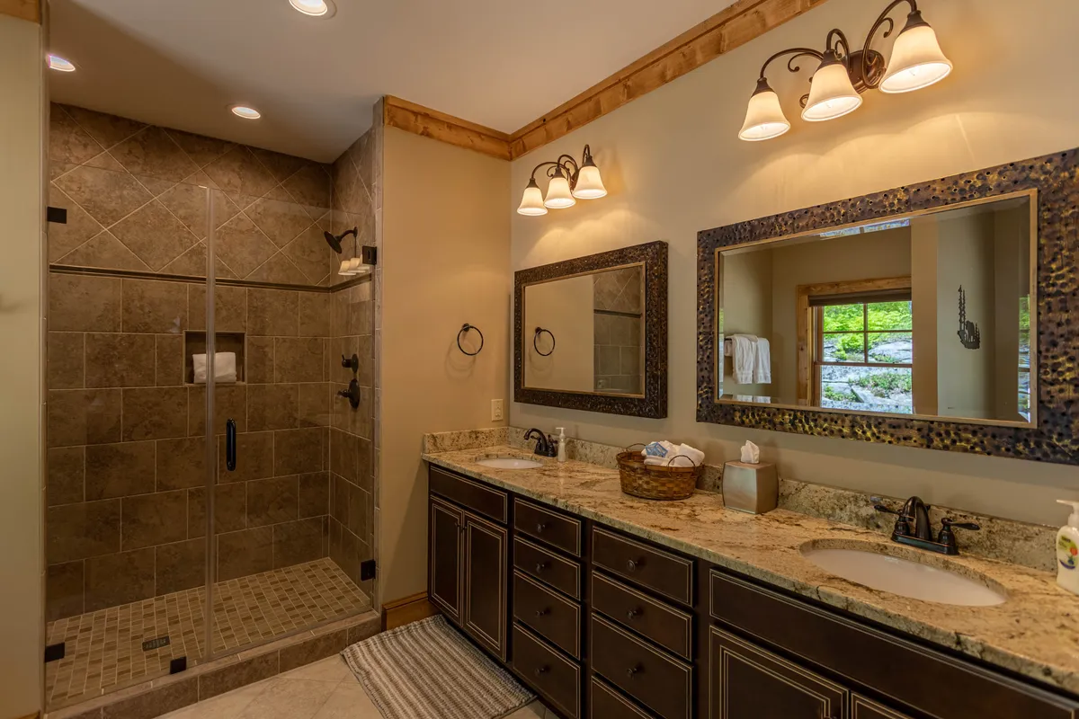 A spa-inspired bathroom featuring a double vanity with a granite countertop, a spacious walk-in shower with custom tile, and large decorative mirrors. Natural light pours in from a window, enhancing the sense of tranquility and luxury.