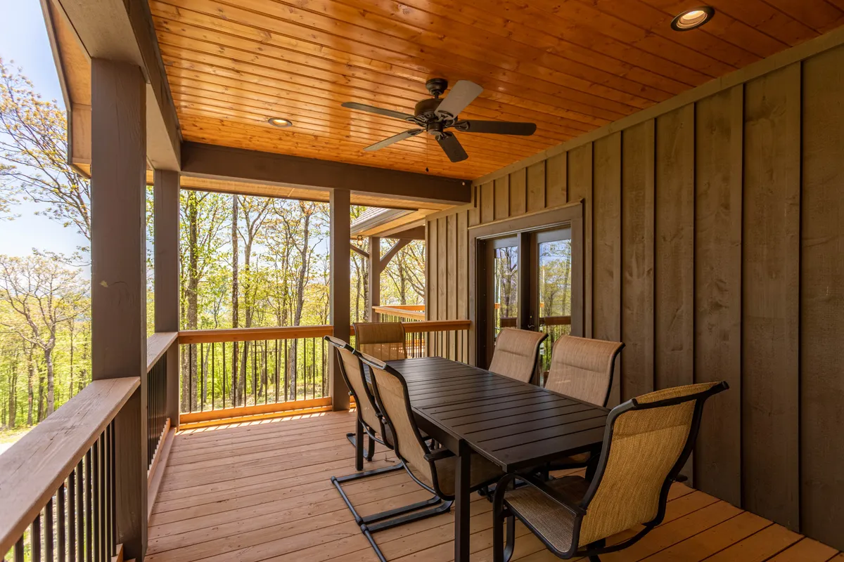 This image features a spacious covered porch with a dining table and chairs, a ceiling fan overhead, wooden railings, and trees encircling the space. The setting is casual and inviting, designed for outdoor dining and gatherings while being shielded by nature.