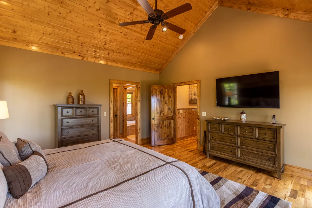 A beautifully furnished bedroom featuring a large bed adorned with decorative pillows, a wooden dresser, a flat-screen TV, and a ceiling fan. Soft natural light filters through the windows, creating a warm, inviting, and serene atmosphere ideal for relaxation.