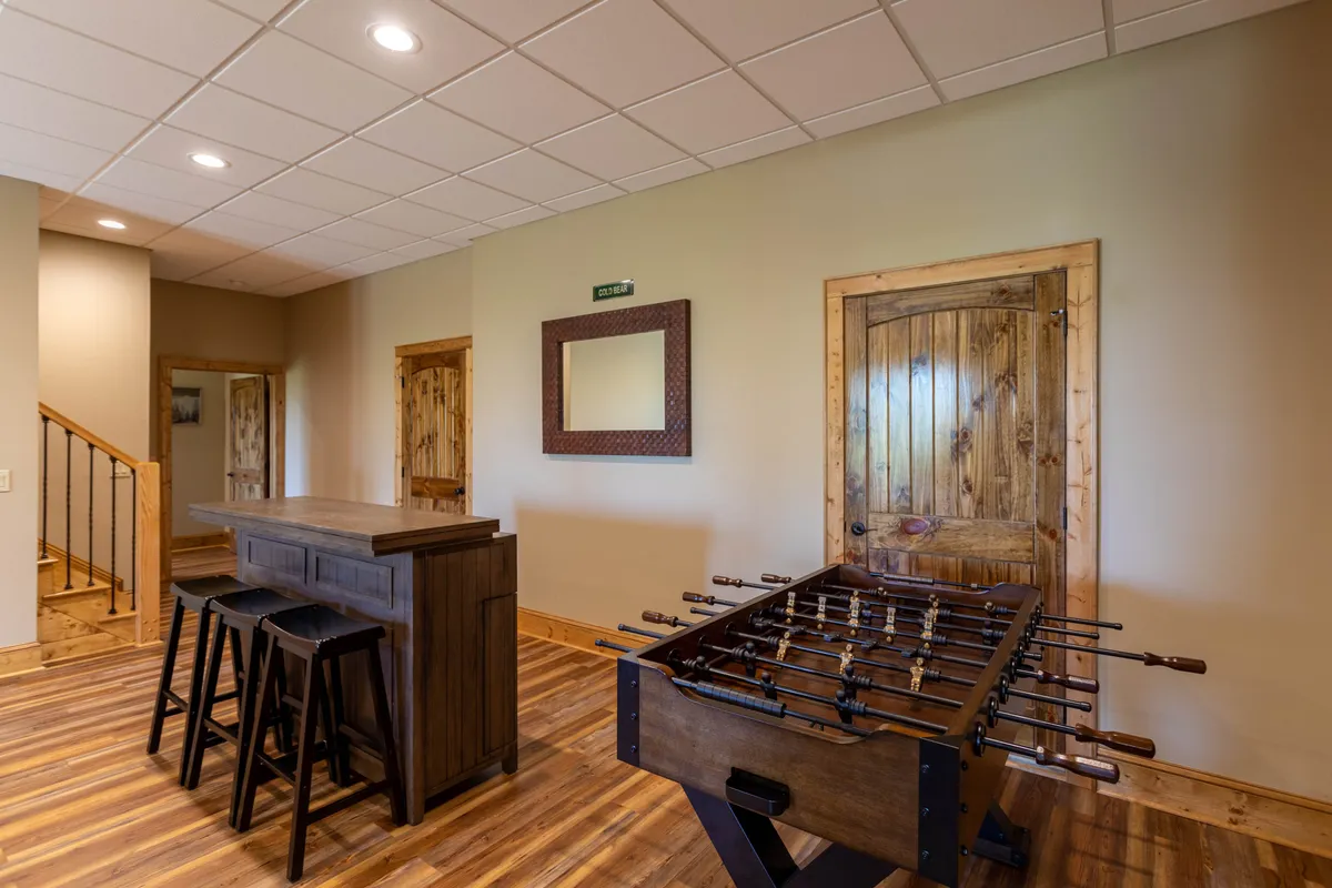 A brightly lit interior spotlights a foosball table and a wooden bar with stools and a mirror in the background. The casual layout evokes a lively, social vibe for friendly competition and gatherings.
