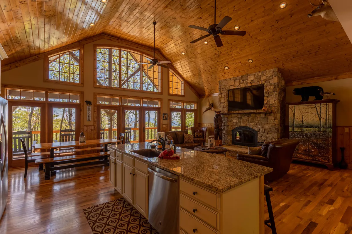 A welcoming interior displaying wide windows with scenic views, a rustic stone fireplace, a granite kitchen island, and a wooden dining table. The open layout connects living, dining, and kitchen spaces, emphasizing communal experiences and relaxation.