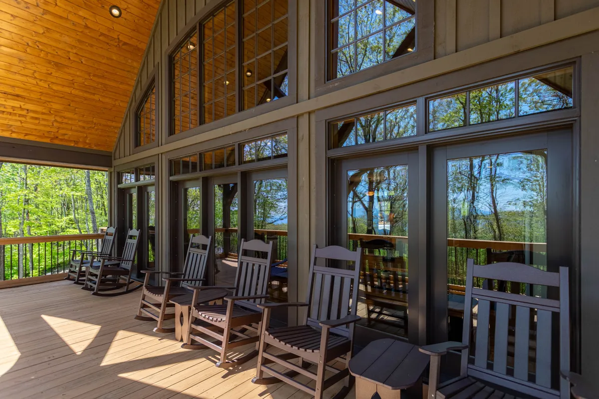 The exterior shows a cozy wooden deck outfitted with rocking chairs and surrounded by lush greenery. Large windows provide a glimpse into the home, and the tranquil porch invites relaxation or wildlife observation after outdoor activities.