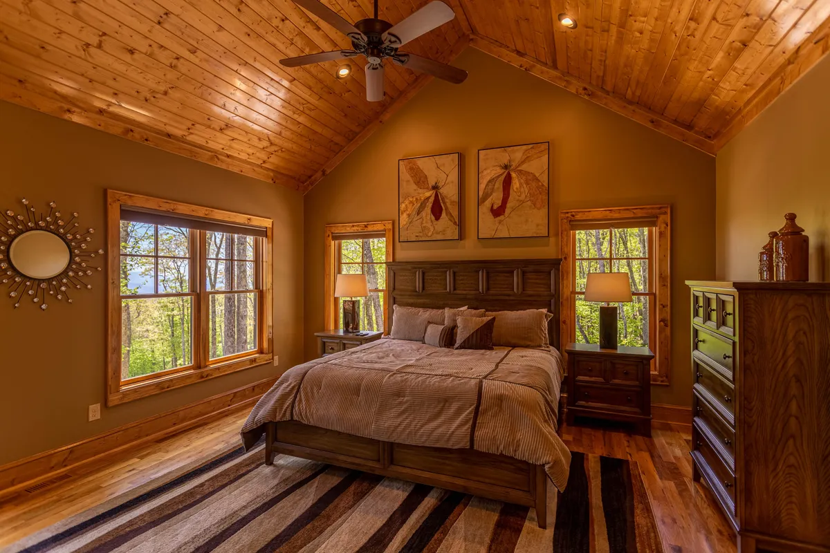 A tranquil bedroom with a large bed, window views of the surrounding trees, tasteful wall art, a dresser, and a mirror. The room exudes a peaceful, inviting ambiance, with abundant natural light streaming in, perfect for unwinding.