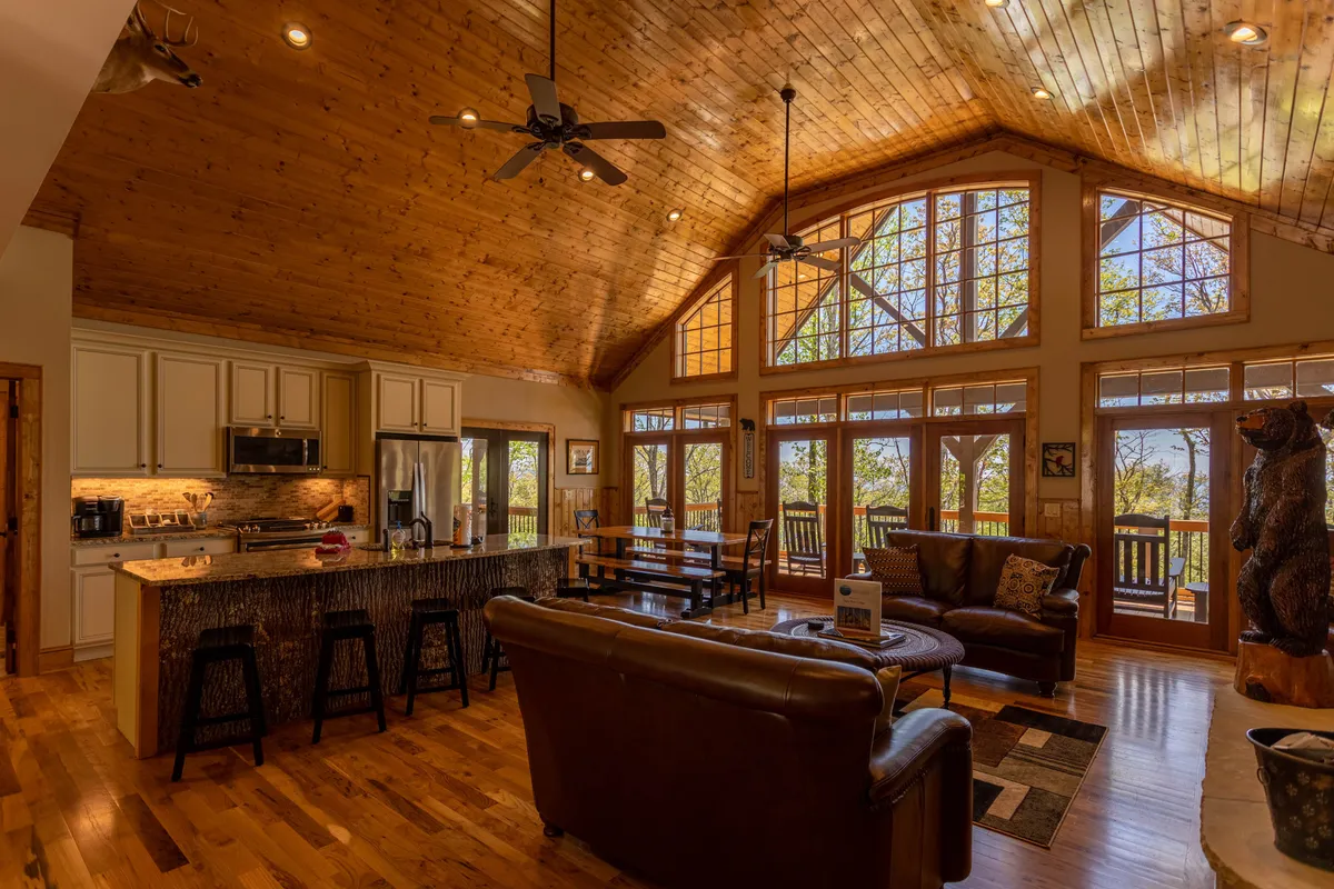 This image features a spacious, open-concept living area with large windows flooding the room with natural light, wooden beams and ceiling, a prominent stone fireplace, comfortable seating, and a kitchen island with bar stools. The setting exudes warmth and invites family gatherings, relaxation, and scenic enjoyment.
